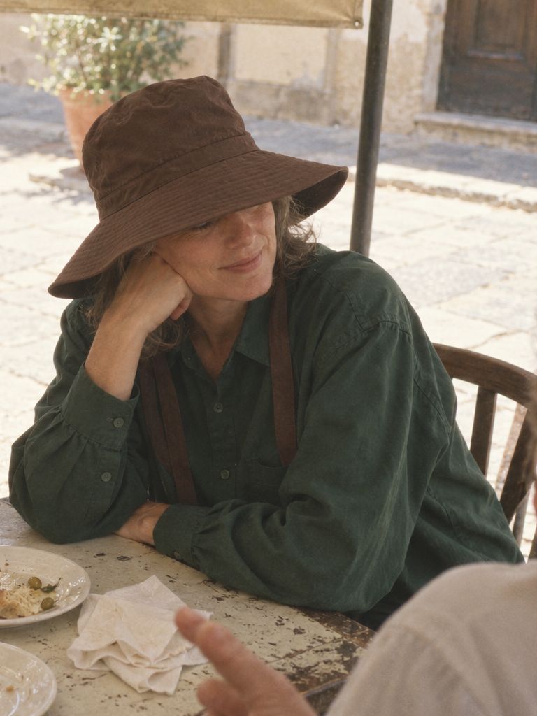 A woman rests her chin on her hand at a long lunch under an awning, in the shade green poplin shirt and the chocolate wide-brim hat