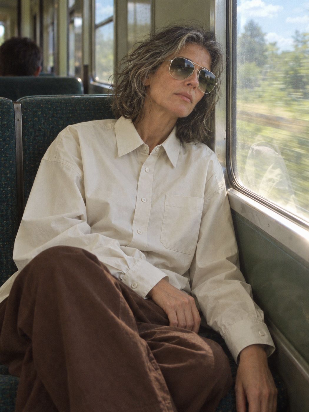 A woman leans on a train window in the bone poplin shirt, the chocolate wide pant and the silver metal aviator