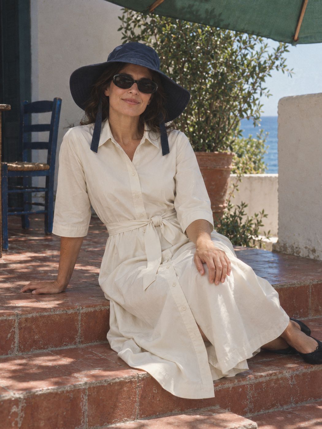 A woman sits on the sunny steps of a seaside terrace in the bone shirt dress, the ink wide-brim hat and the ink chunky oval